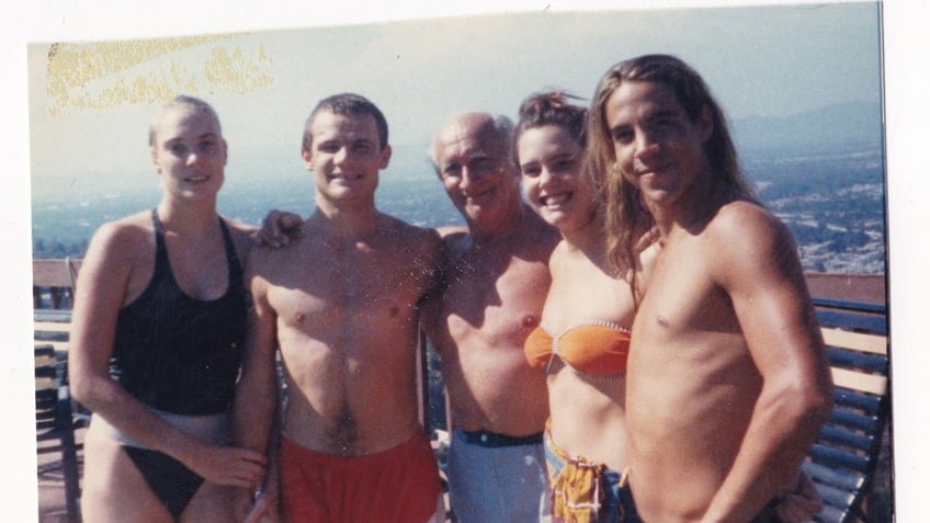 Ione Skye in an orange printed bikini posing with Anthony Kiedis in shorts and other people around them smiling.