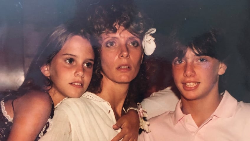 A young Ione Skye holding on to her mother who is wearing a white dress and a flower in her hair as she holds her son.