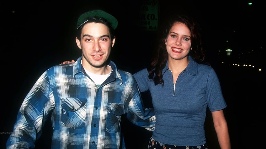 Adam Horovitz wearing a blue plaid shirt with his arm around Ione Skye who is wearing a blue shirt