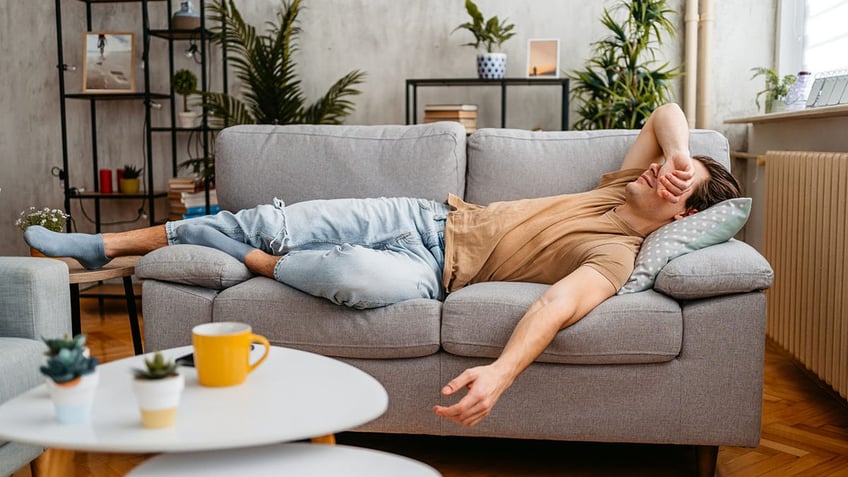 Man laying on a gray couch. The man is wearing blue jeans and a tan top.