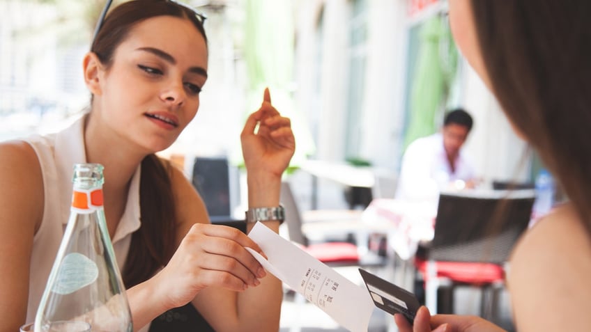 A woman passes a receipt from a meal at a restaurant to another woman, who has her credit card in her hand.