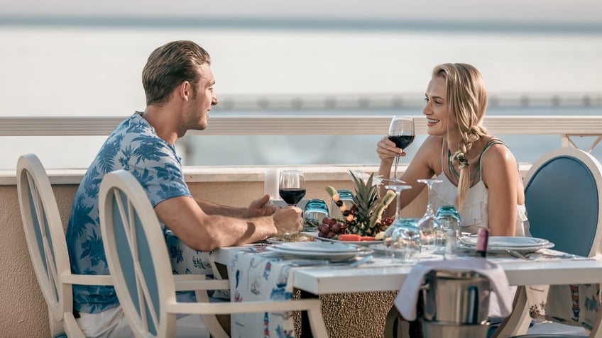 A young couple drink red wine while dining outside along the waterfront.