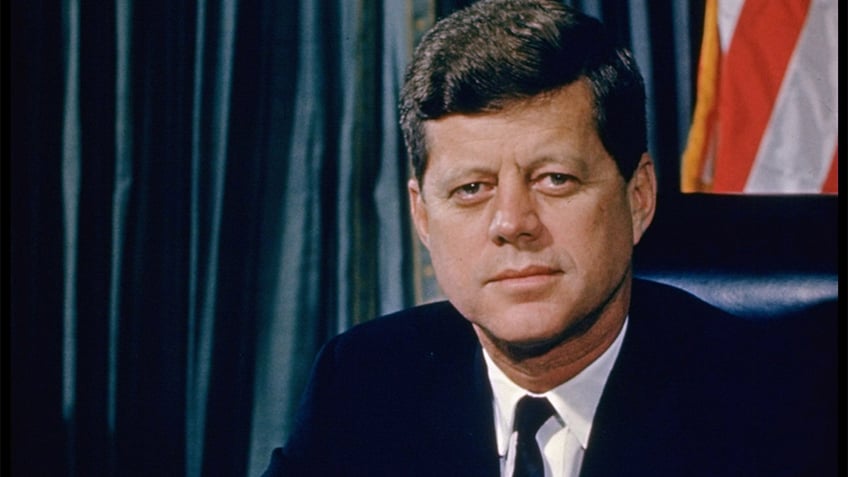 President John F. Kennedy posing for a picture at his desk with a US flag in the backdrop.