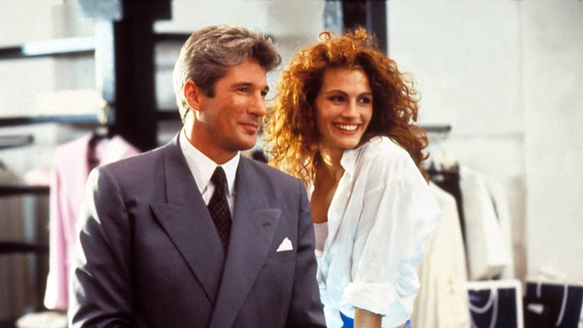 Richard Gere in a dark grey suit soft smiles next to Julia Roberts in a white shirt and blue skirt in character during "Pretty Woman"