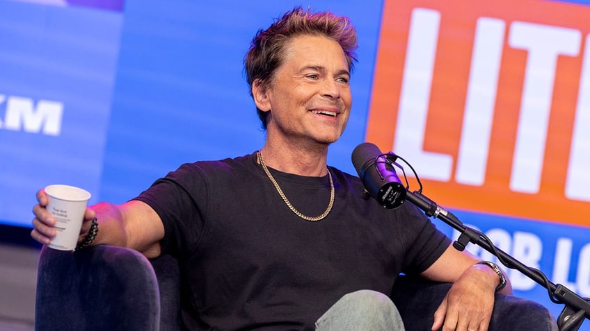 Rob Lowe sits in a chair wearing a black shirt and gold chain taping his podcast