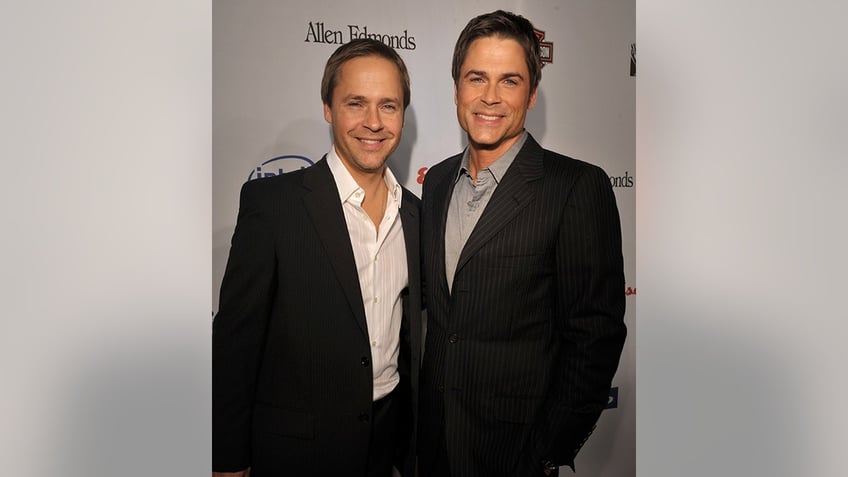 Chad Lowe in a black suit and white button down smiles next to his big brother Rob Lowe in a black suit and grey shirt