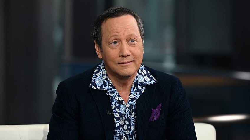 Rob Schneider in a dark navy jacket and printed blue shirt sitting on a chair