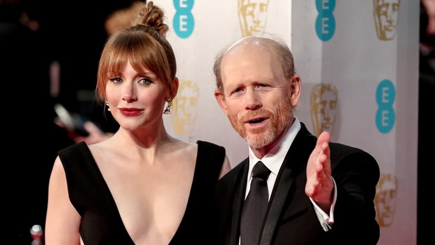 Bryce Dallas Howard in a bun and low plunging black dress next to father Ron Howard who holds his hand out towards the camera