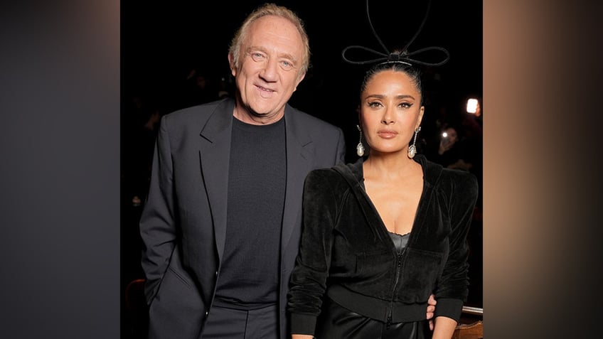 François-Henri Pinault in a grey suit and black shirt smiles next to wife Salma Hayek in a black dress and velour jacket at the Balenciaga RTW Spring show