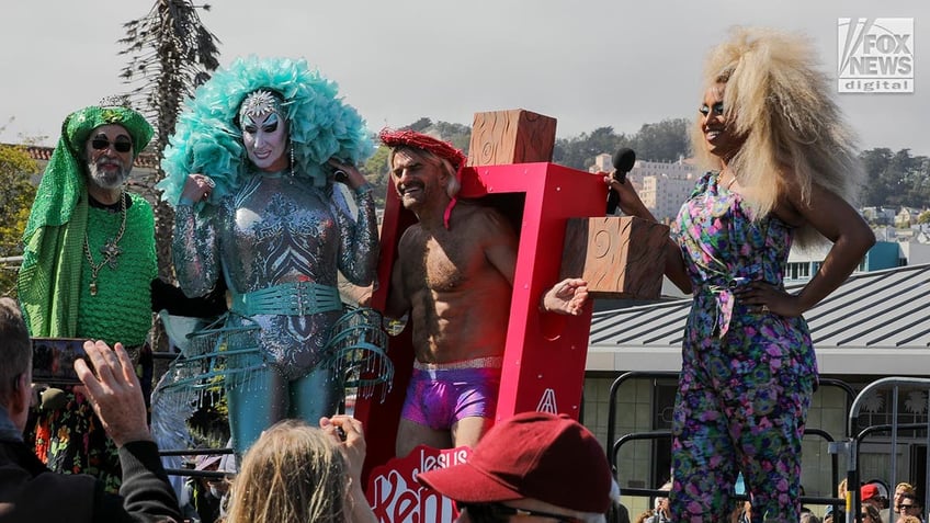 A man in an oversized box simulating a Ken doll box with "Jesus Ken" written on it and a cross coming from its sides.