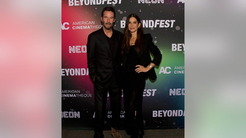 Keanu Reeves and Sandra Bullock, both in black outfits, hold onto each other on the carpet