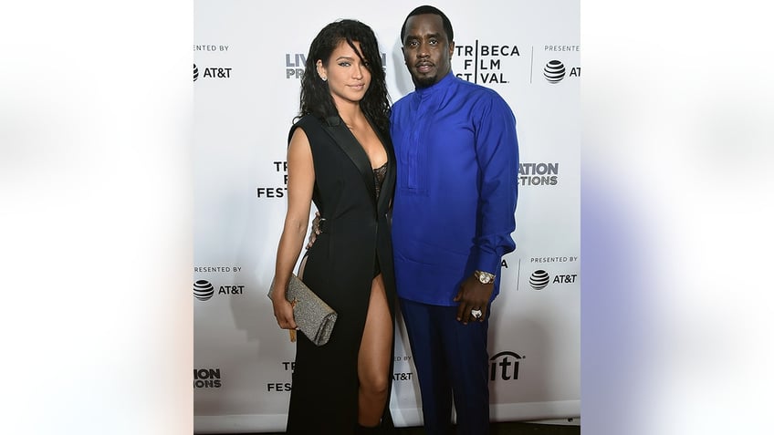 Cassie in a long black dress with a slit stands next to Diddy in a royal blue shirt on the carpet
