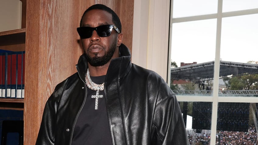 Diddy looks serious in a leather jacket and black shirt with a diamond cross chain and black sunglasses
