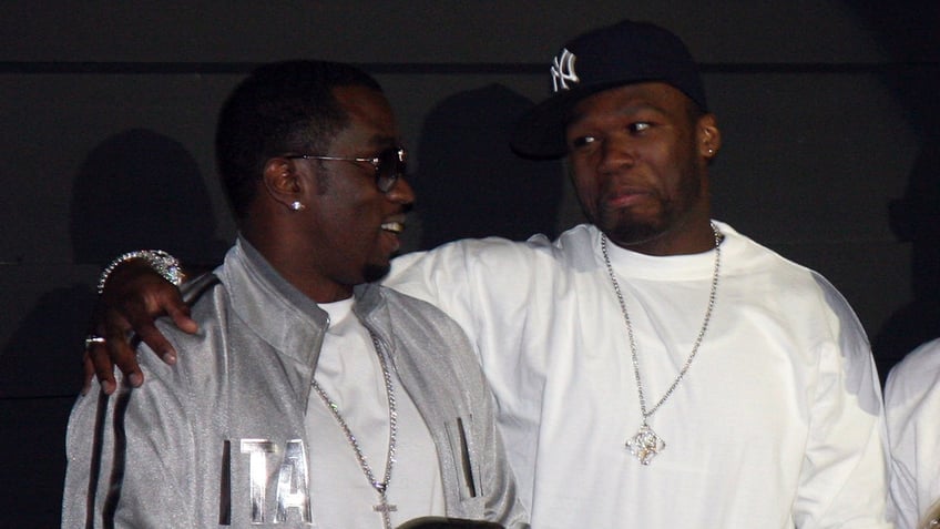 50 Cent in a Yankees fhat and white shirt wraps his arm around P Diddy in a silver jacket and shades in Las Vegas