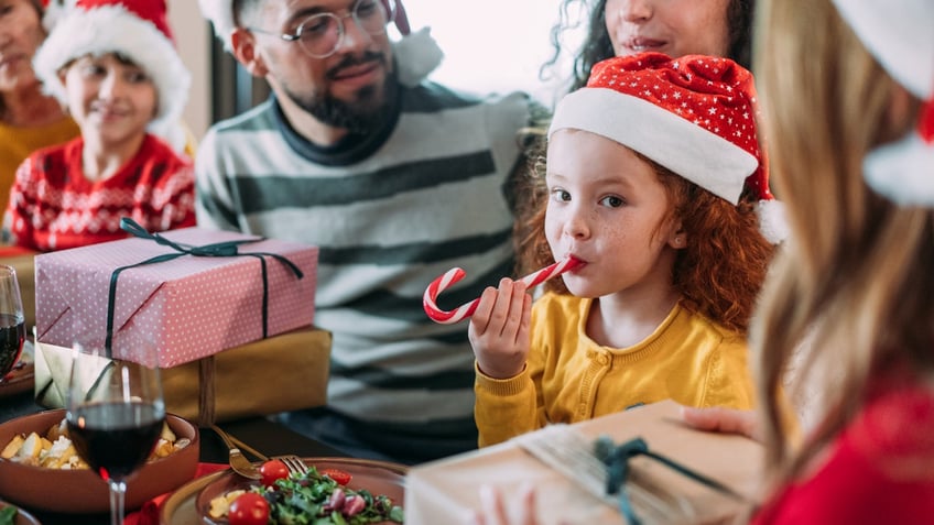 A girl eats a candy cane from the bottom while staring into the camera.