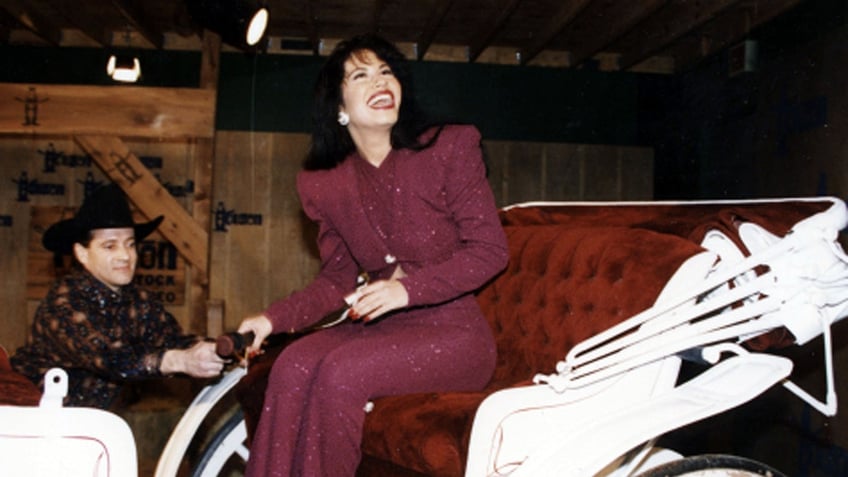 Selena Quintanilla in a maroon pant suit looks up and laughs while sitting in a horse drawn carriage