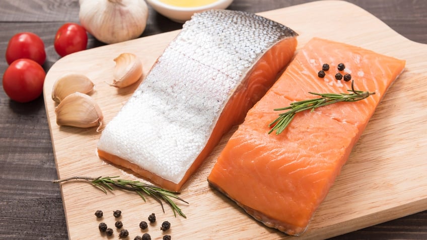 Two filets of salmon, one with the skin facing up, rest on a wooden cutting board with onions, basil and pepper.
