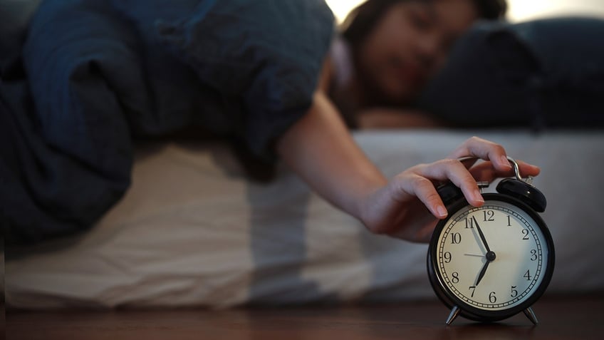 A young Asian woman tries to turn off the alarm clock in her bedroom at home while still lying in bed.