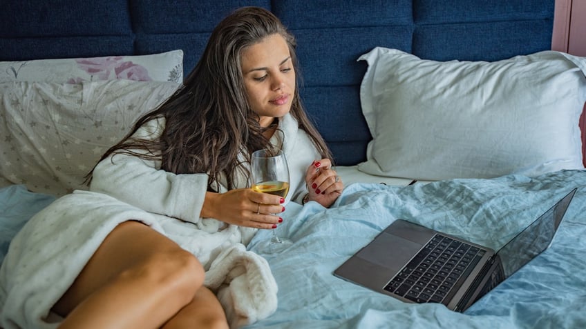 A woman drinking white wine in bed looks at her laptop.