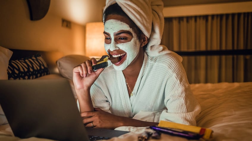 A woman with a night mask on her face eats a chocolate bar in bed while looking at her laptop.