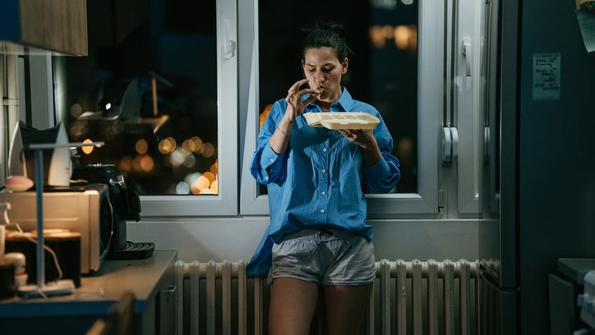 Woman standing in kitchen eating late at night