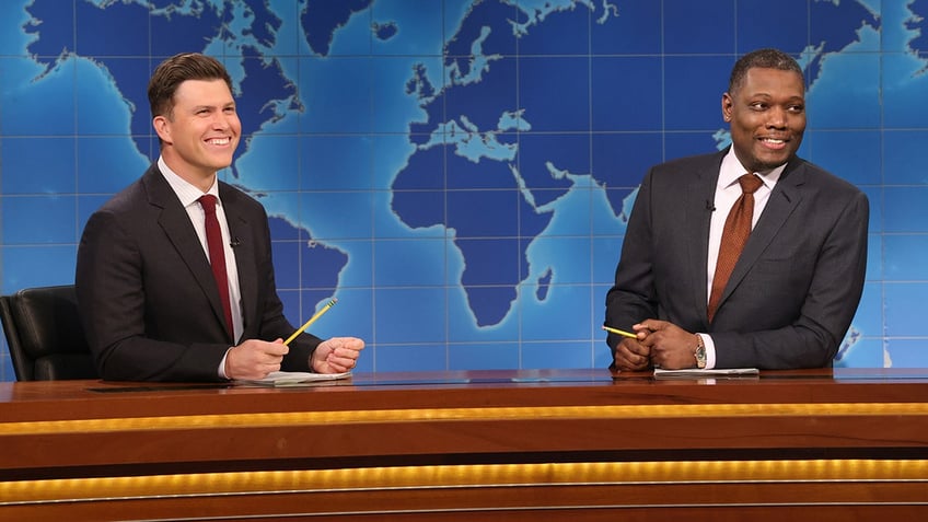 Colin Jost and Michael Che sit behind the desk during 'Weekend Update' on 'SNL' wearing dark jackets and a red an orange tie, respectively,