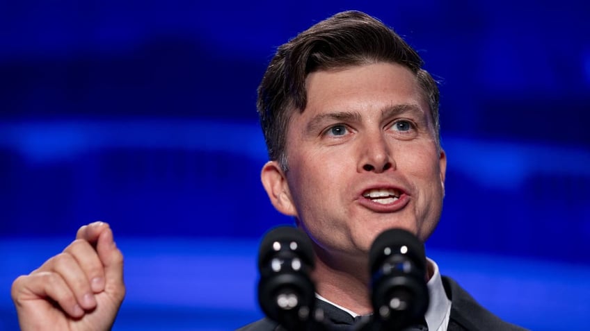 Comedian speaking at podium on stage with microphone, blue background.