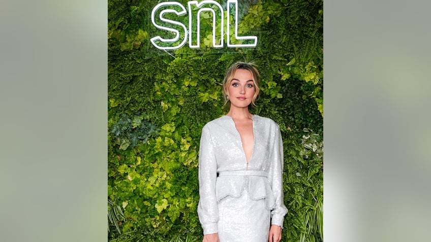 Chloe Fineman in a sparkly two piece blazer and skirt looks directly at the camera, in front of foliage and a sign that says 'SNL'