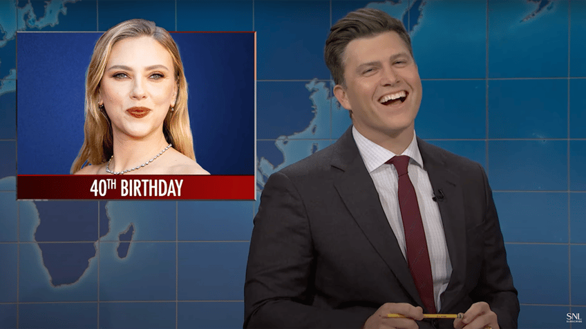 Colin Jost laughs in a grey suit and red tie behind the "SNL" 'Weekend Update" desk inset a photo of Scarlett Johansson that says "40th birthday'