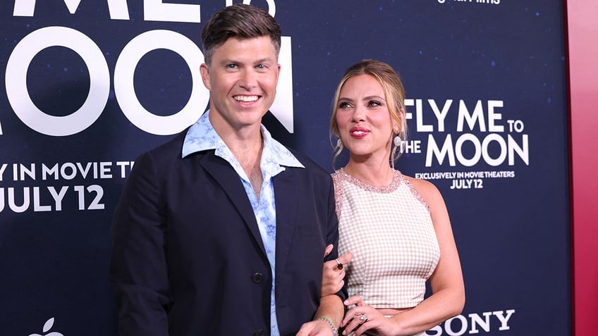 Scarlett Johansson in a white dress holds on to husband Colin Jost, in a navy suit and blue shirt, and looks at him lovingly on the carpet