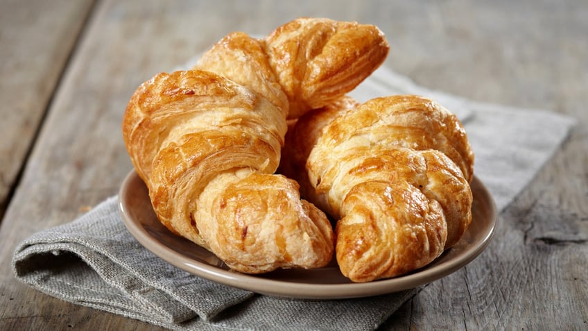 Two butter croissants rest on a plate beneath a cloth napkin on a wooden table.