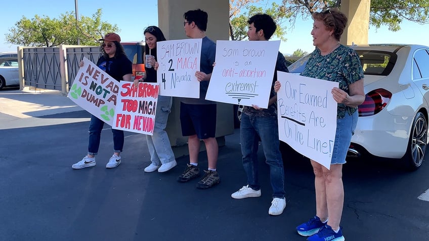 Protesters hold signs criticizing GOP Senate hopeful Sam Brown