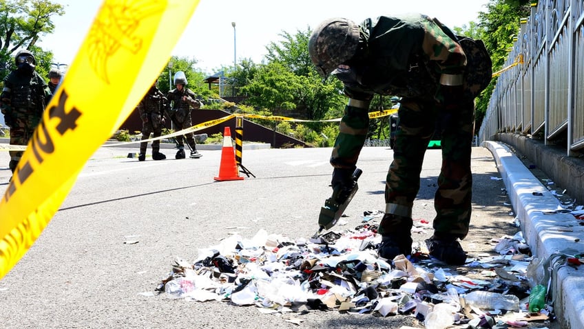 A South Korean soldier wearing protective gears checks the trash from a balloon, presumably sent by North Korea, behind yellow crime scene tape.