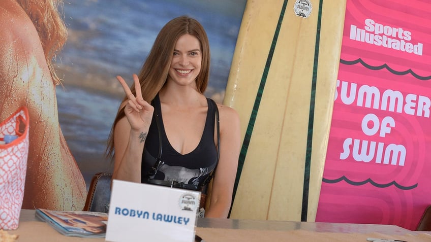 Robyn Lawley in a black one-piece bathing suit throws up a piece sign as she smiles for a photo