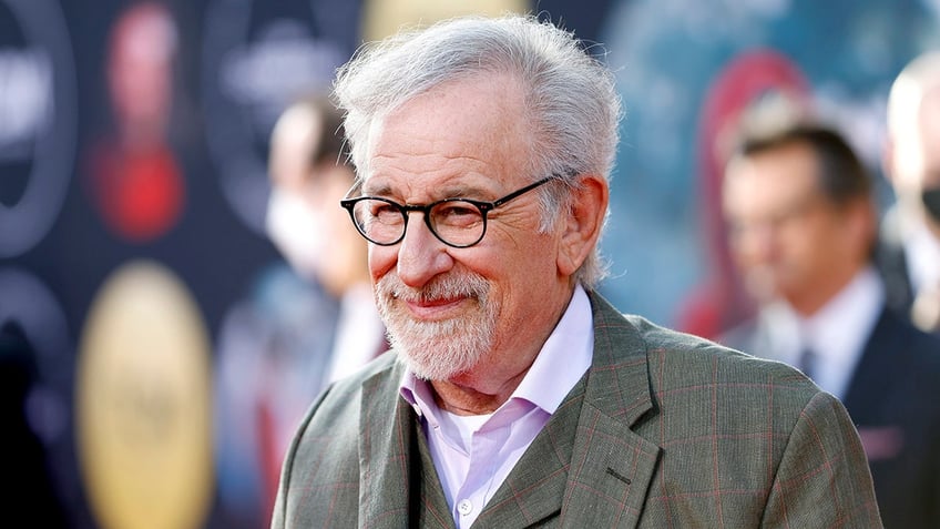 Steven Spielberg in a grey plaid jacket and black rimmed glasses smiles while walking the carpet