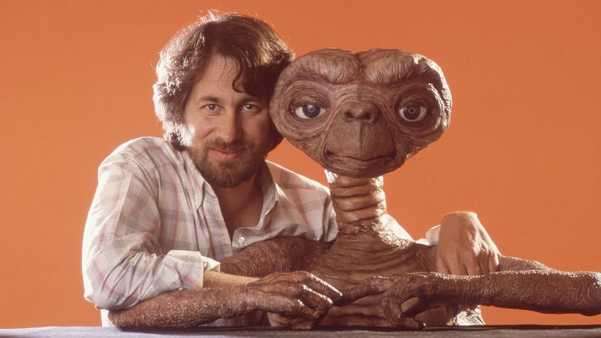 Steven Spielberg in a plaid shirt smiles with E.T. in front of an orange back drop