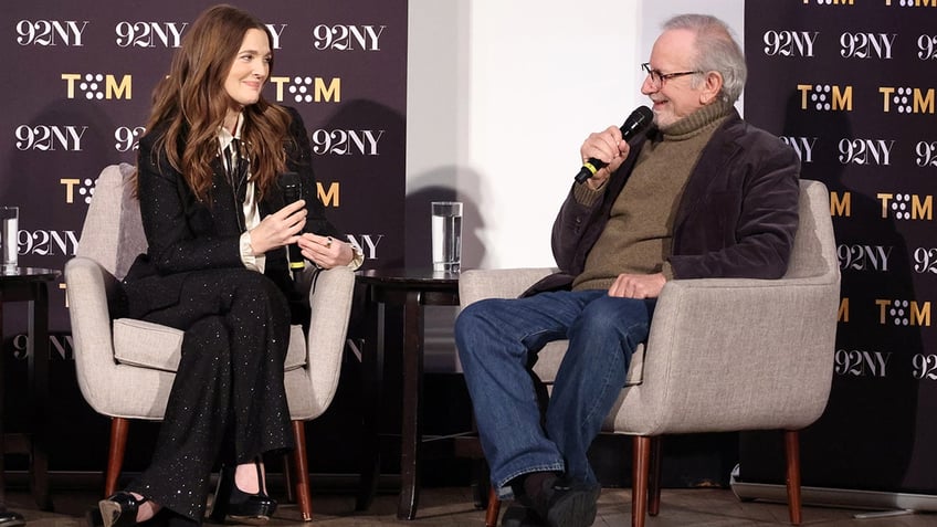 Drew Barrymore in a sparkly black pantsuit soft smiles on stage at Steve Spielberg sitting opposite her in blue jeans and a velvet brown suit