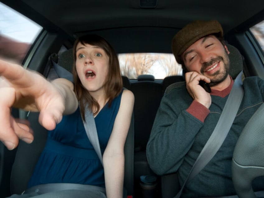 A young woman sees an accident coming, while the man, who is driving, is talking in his ce