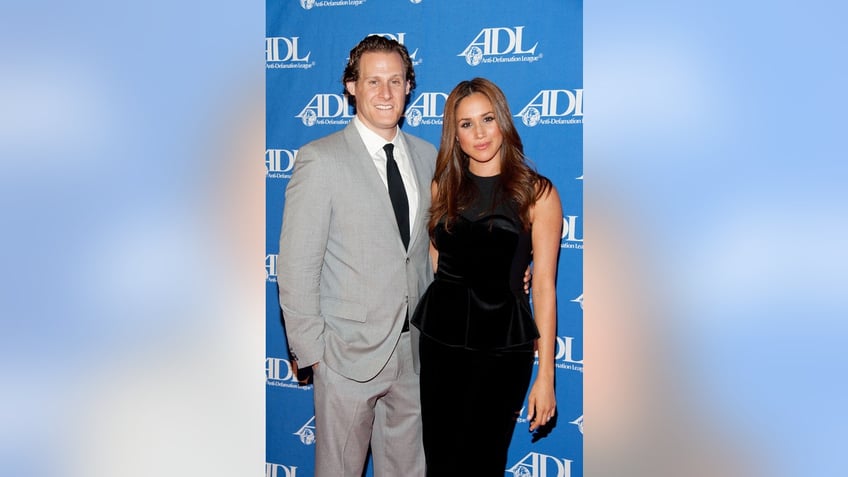 Meghan Markle in a black sleeveless dress standing next to Trevor Engelson in a grey suit..