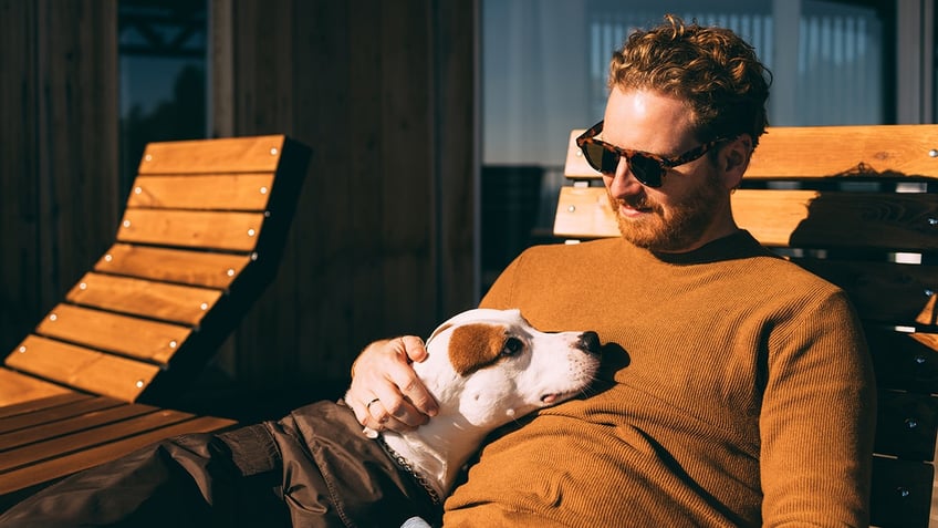 Man in sunglasses bonding with dog on a wooden bench.