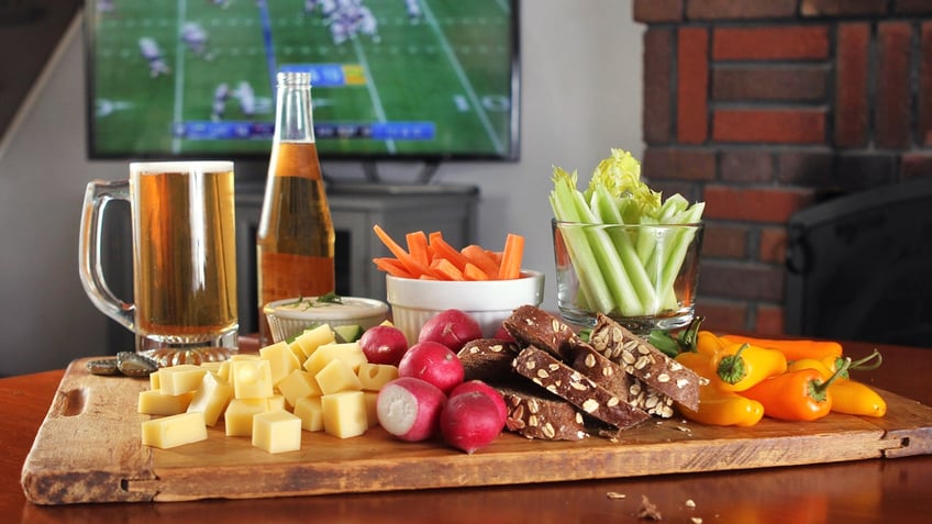 A football game is playing on TV in the background. In the foreground are healthy party food options and some beer.