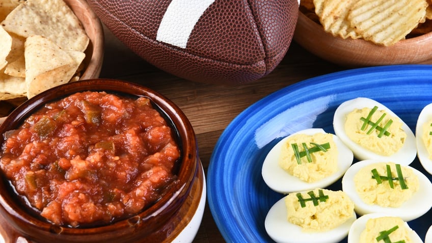 Deviled eggs decorated to look like footballs are shown on a plate on a table next to a football and chips and salsa.