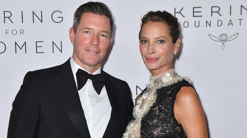 Ed Burns in a classic tuxedo soft smiles on the carpet with wife Christy Turlington in a black lace dress