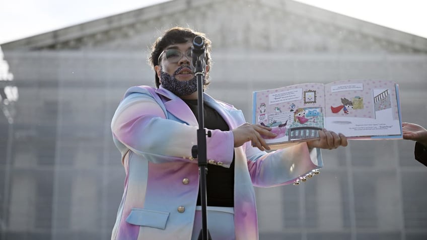 A speaker in drag wearing a pastel rainbow suit reads a children’s book aloud at a microphone during a public event outside the U.S. Supreme Court