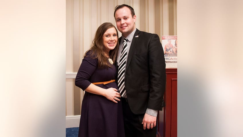 Anna Duggar wearing a dark purple dress standing next to her husband Josh Duggar in a dark suit and striped tie