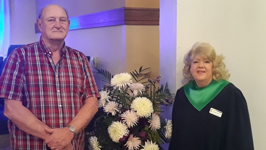 Barbara Mabee Abel wearing a formal graduation gown standing next to her husband Jim in a plaid shirt