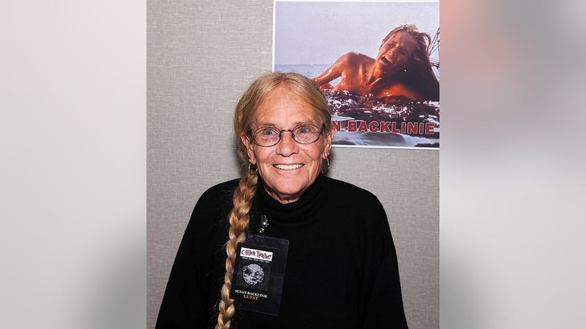 Susan Backlinie in a black top smiles for a photo with her hair in a braid