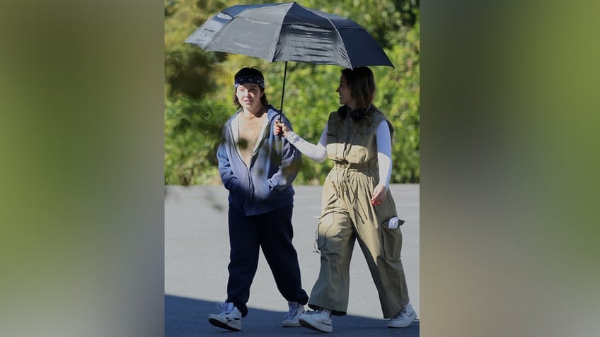 Sydney Sweeney in sweatpants walks on set with someone holding a black umbrella over her