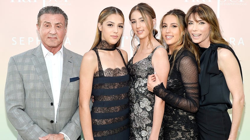 Sylvester Stallone in a grey suit and white shirt stands next to daughters Sistine in a black frill and lace dress, Scarlet in a black lace dress, Sophia in a black sheer dress, and wife Jennifer in a black dress
