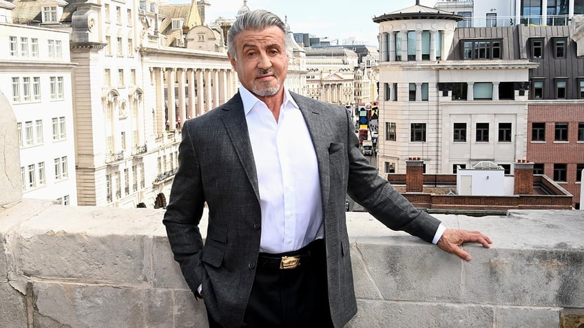 Sylvester Stallone in a dark suit and black pants poses for a photo